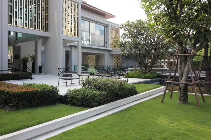 Modern two-story house with large glass windows, surrounded by landscaped gardens and trees, featuring outdoor seating on a patio.