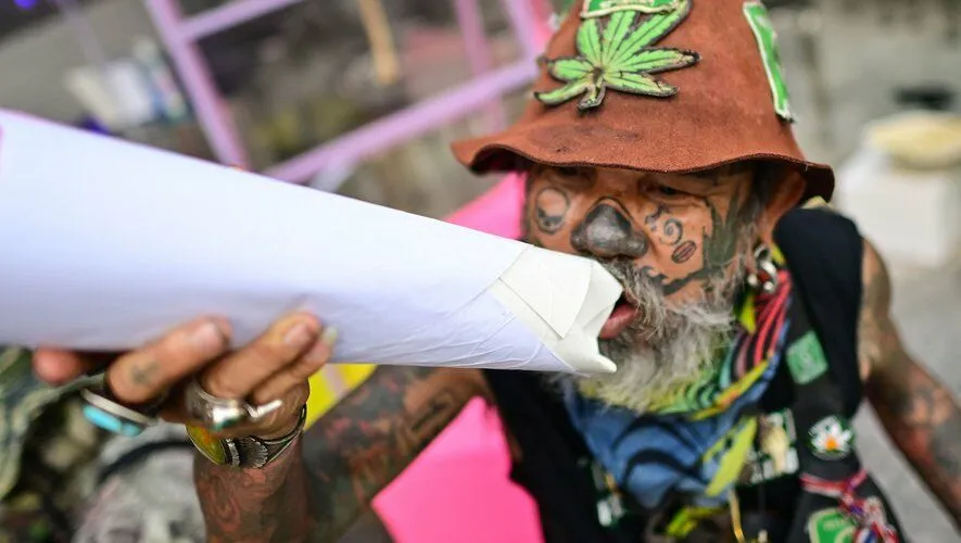 article image - A person with tattoos and a hat decorated with a cannabis leaf holds a large rolled-up paper, appearing as if smoking. A person with tattoos and a hat decorated with a cannabis leaf holds a large rolled-up paper, appearing as if smoking.
