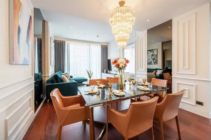 Dining area with a glass table, eight leather chairs, and an elegant chandelier. The space connects to a living room with a sofa and large windows.