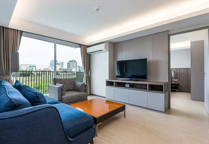 Modern living room with blue sofa, orange coffee table, and TV. Large window with city view and adjacent open bedroom door.