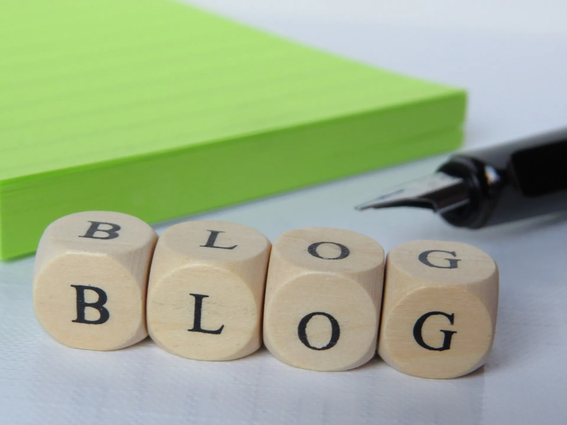 Wooden dice spelling "BLOG" beside a fountain pen and green notepad.