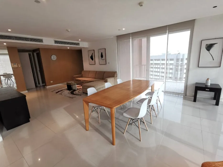 Spacious modern living room with a wooden dining table, white chairs, beige sofa, abstract wall art, and floor-to-ceiling windows.