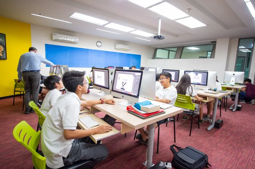 Students in a computer lab work on desktop computers. They are engaged in digital design tasks, seated at desks with a teacher assisting them. The room has colorful walls and bright lighting.
