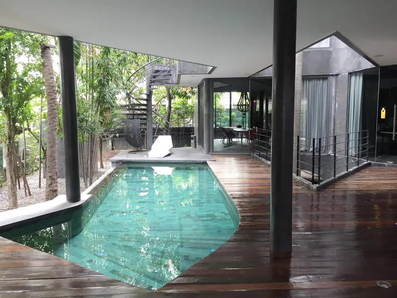 Modern indoor space with a sleek pool, surrounded by wooden flooring and columns, leading to a glass-walled room and a spiral staircase, with greenery outside.