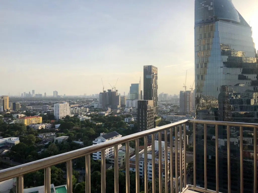 City skyline view from a balcony showing high-rise buildings, cranes, and a glass-covered tower reflecting the sunset.