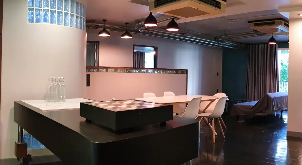 Modern interior with a dining table and chairs, pendant lights, and a chessboard on a side table, featuring glass block walls and exposed ductwork.