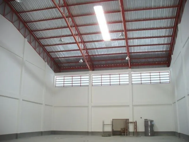 Interior view of a spacious, empty warehouse with high ceilings, red beams, and fluorescent lighting.