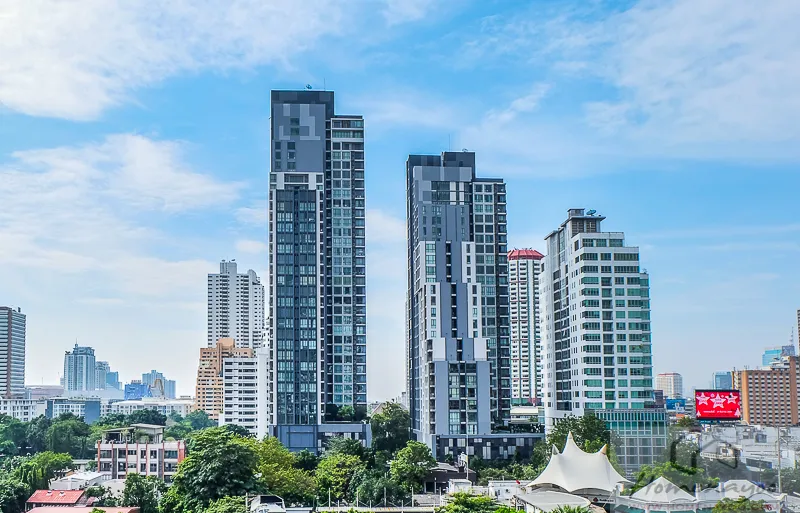 Modern cityscape with tall skyscrapers under a clear blue sky, surrounded by smaller buildings and greenery.