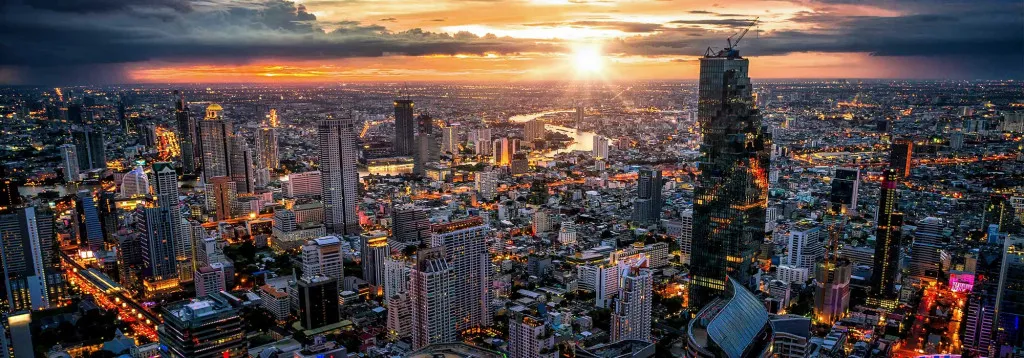 Aerial view of a city skyline at sunset, featuring illuminated skyscrapers and a vibrant river reflecting the colorful sky.