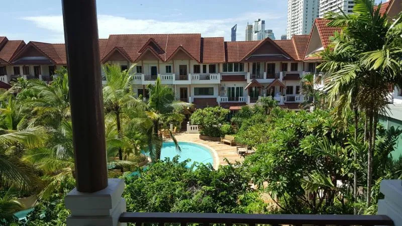 View of a lush courtyard with a pool surrounded by tropical plants, overlooked by a building with red-tiled roofs and balconies, against a backdrop of city skyscrapers.