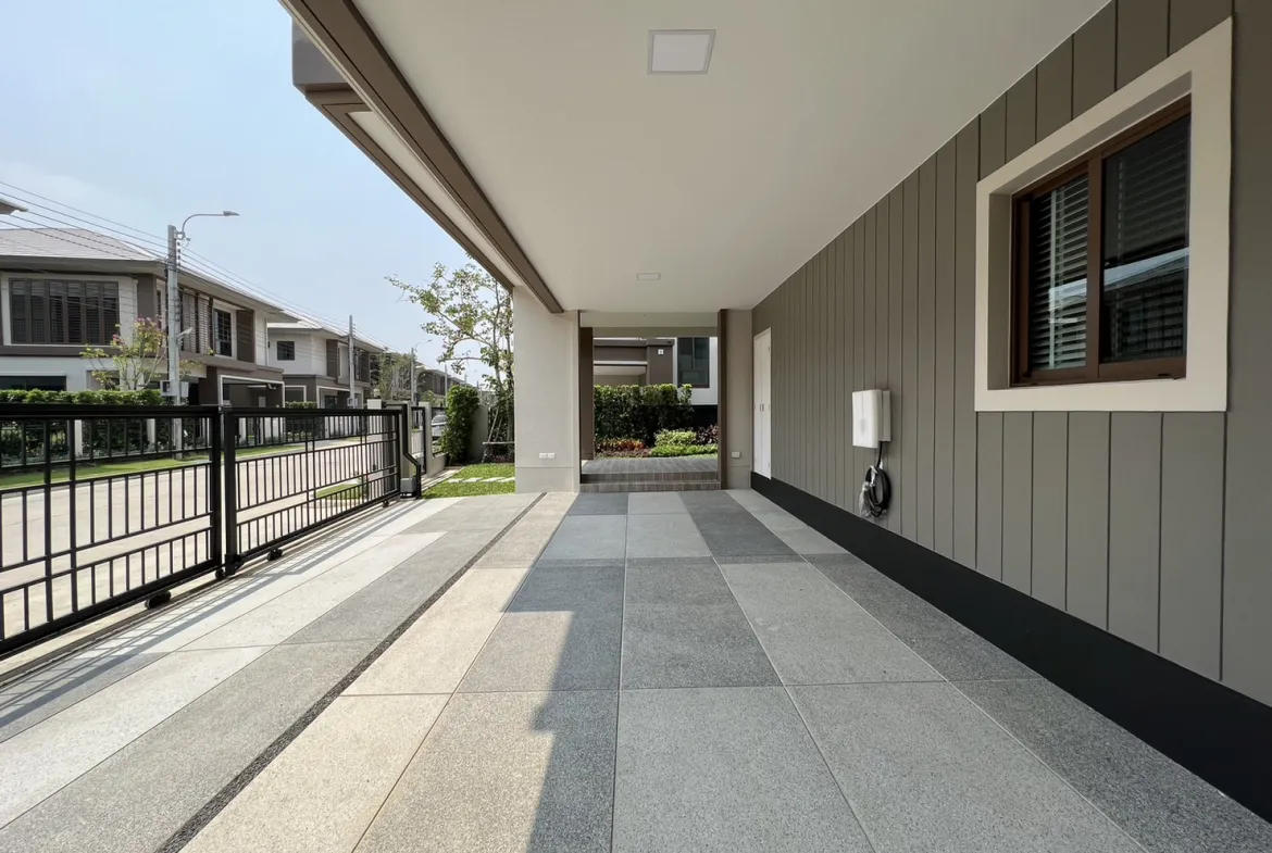 Covered driveway and entryway of a modern home, featuring a paved floor, exterior walls, a large window, and a secured gate, with neighboring houses visible across the street.