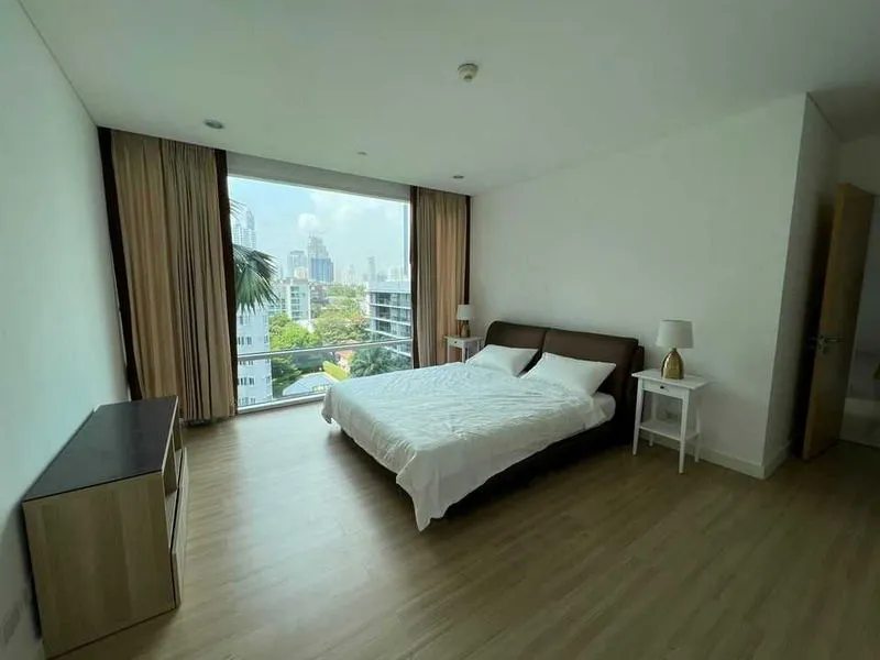 Spacious bedroom with a large bed, wooden floor, nightstands with lamps, and a view of a cityscape through a floor-to-ceiling window with curtains.