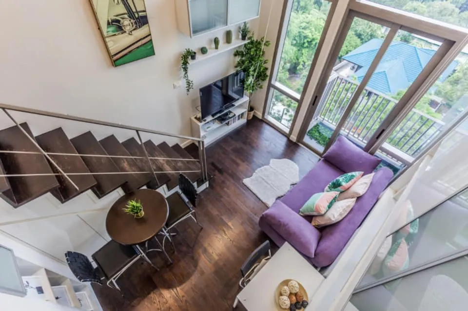 A modern loft apartment with a purple sofa, TV, dining table, and large windows overlooking greenery, featuring dark wood flooring and stairs.