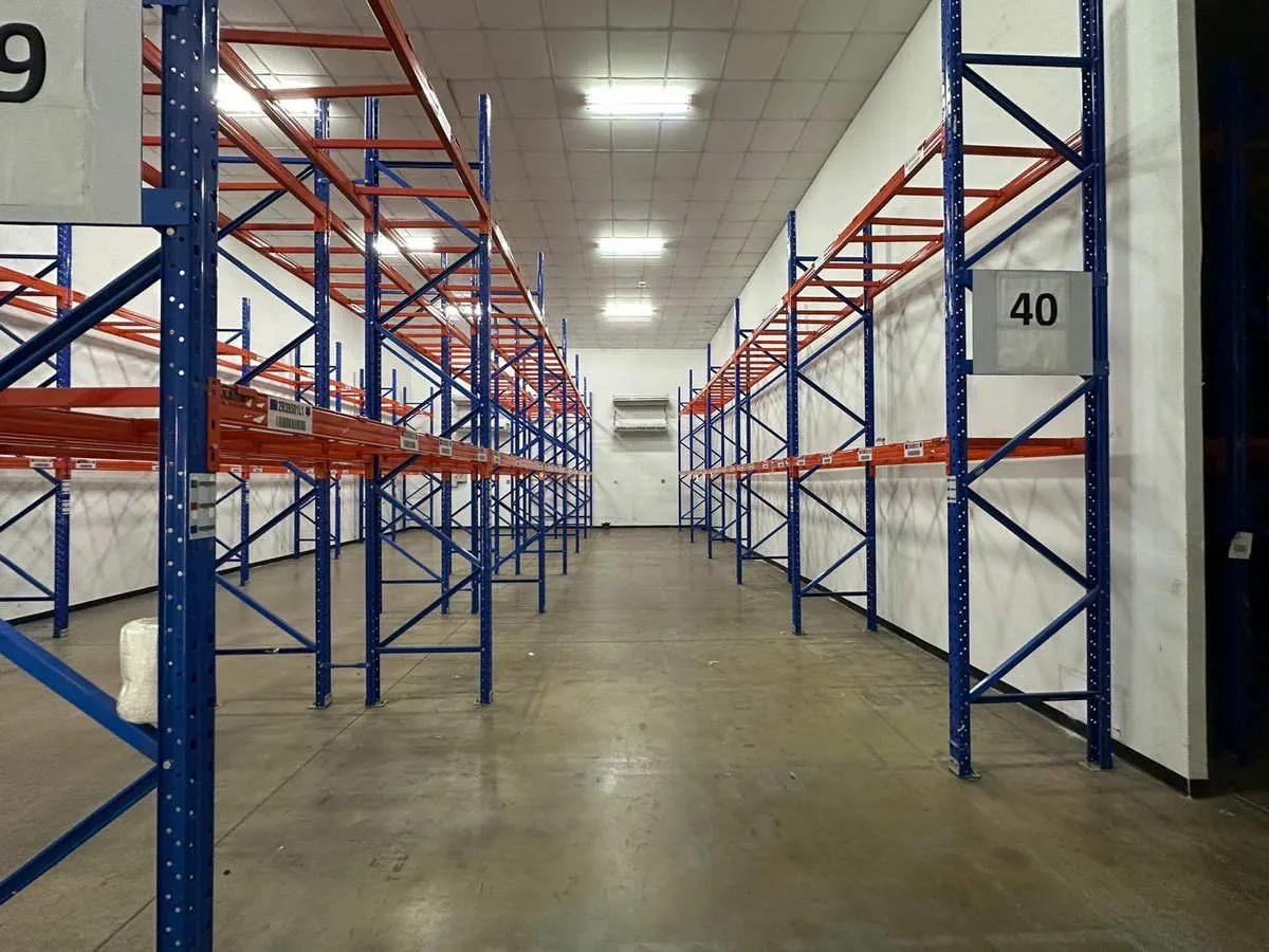 ทางเดินในโกดังว่างเปล่า มีชั้นวางของโลหะสีฟ้าและสีส้ม หมายเลข 39 และ 40 วางอยู่ใต้แสงไฟฟลู