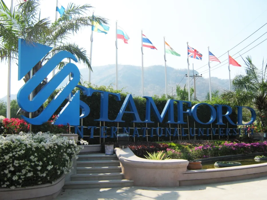 Stamford International University entrance sign with flags of various countries in the background, surrounded by palm trees and flowers.