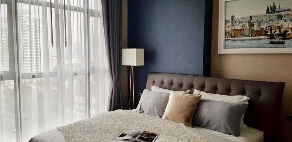 Bright bedroom with large window, gray tufted headboard, and blue accent wall, featuring a cityscape painting and a floor lamp.