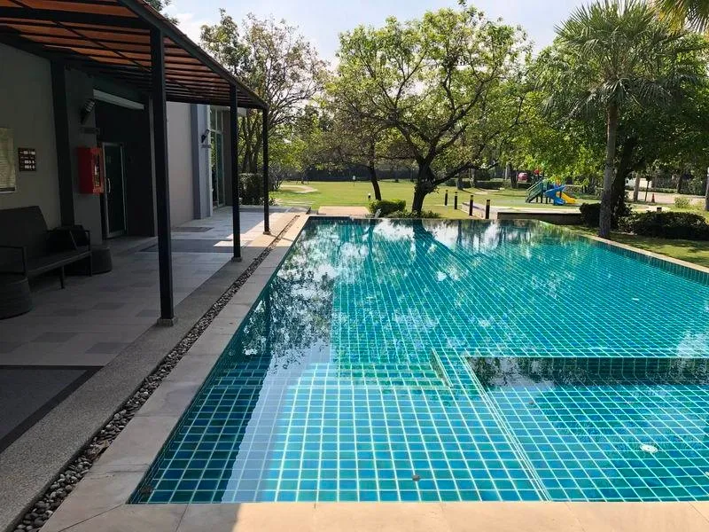 Outdoor swimming pool with blue tiles next to a building, surrounded by greenery and a play area in the background.