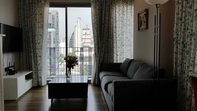 A modern living room with a gray sofa, floor lamp, and a glass coffee table with flowers. Large window with city view and patterned curtains.