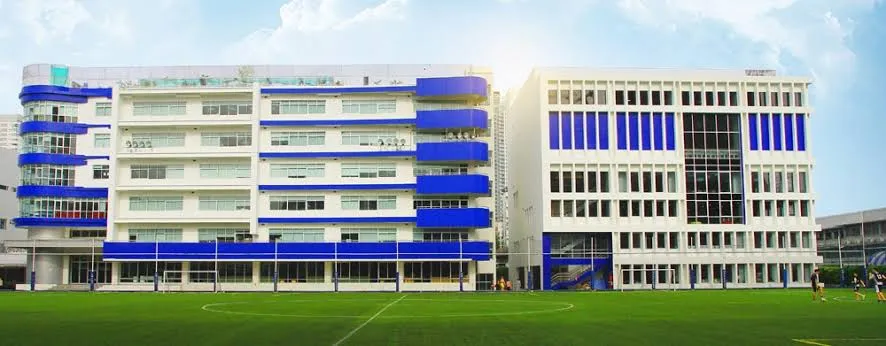 A modern school building with blue and white accents, featuring multiple stories and large windows, set next to a green sports field under a cloudy sky.