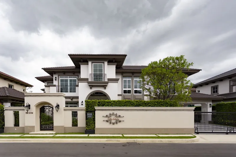 Two-story modern house with gray roof, large windows, and arched entryway, surrounded by a white wall and greenery under a cloudy sky.