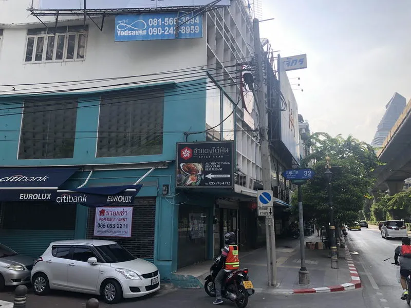 Street scene with a blue and white building featuring a Thai restaurant sign, parked cars, and a person on a motorcycle.