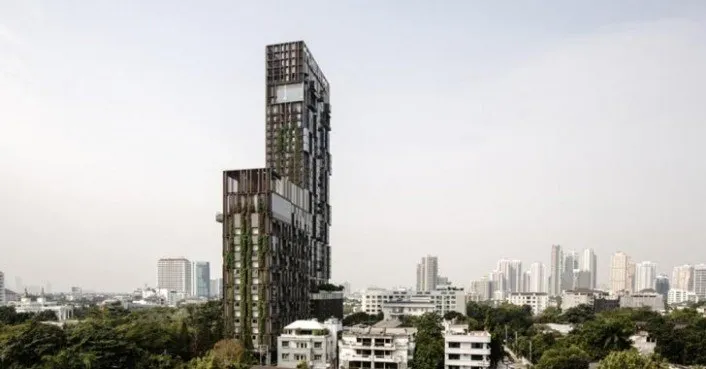 A unique, modern skyscraper with a mix of glass and green plant facades, set against a cityscape with other buildings and a clear sky.