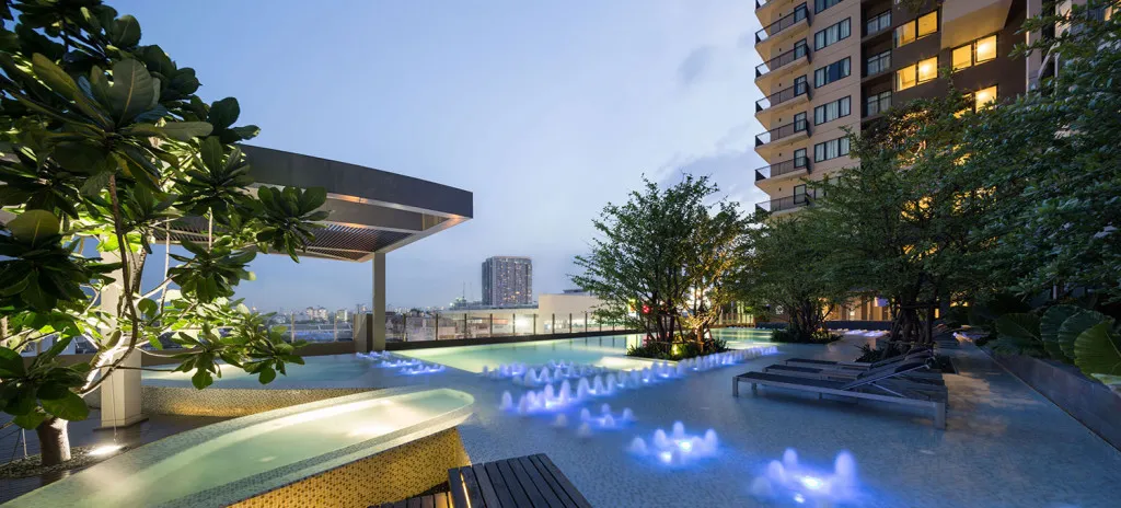 Rooftop pool with blue lights, surrounded by trees and sun loungers, adjacent to a modern high-rise building at dusk.