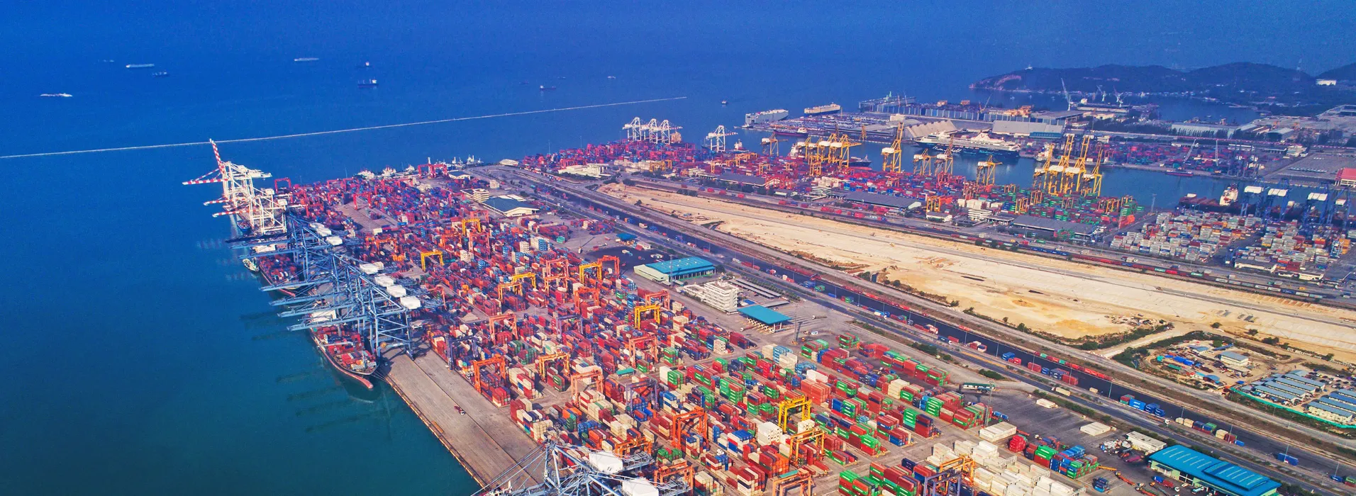 Aerial view of a busy shipping port with colorful cargo containers, cranes, and docked ships by the ocean.