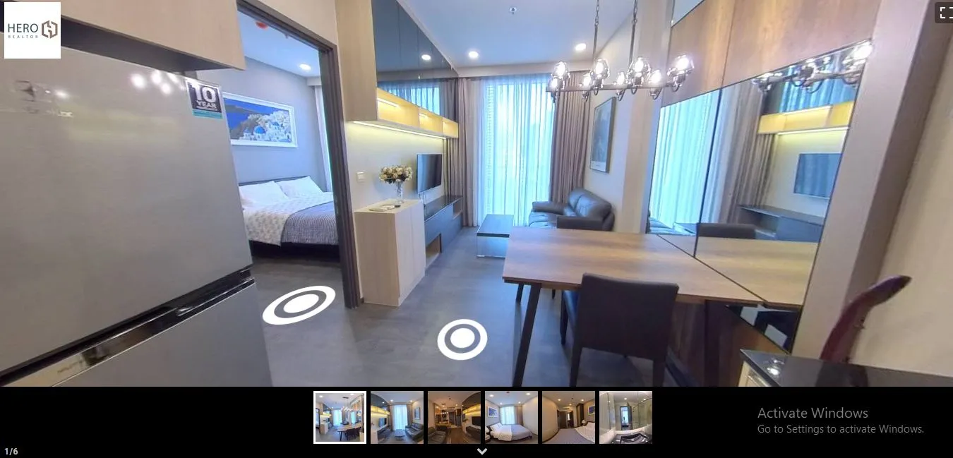 Modern apartment interior with open concept living and dining area, featuring a wooden dining table, leather sofa, and wall-mounted TV. Visible bedroom and decorative chandelier.