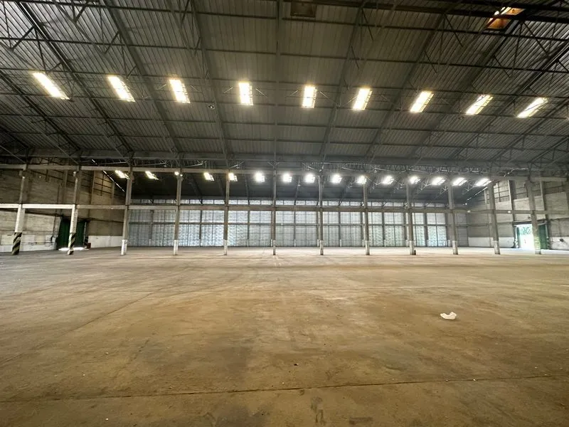 Spacious empty warehouse interior with high ceiling, exposed steel beams, and bright overhead lights creating an industrial atmosphere.