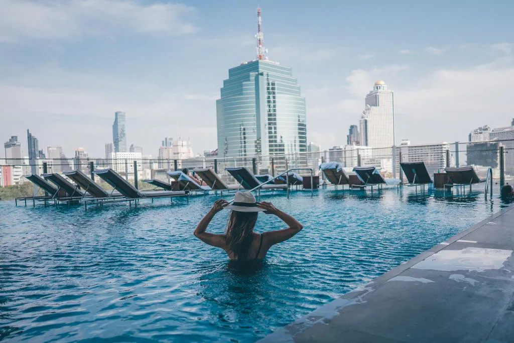 Woman in a pool wearing a hat, looking at a skyline with tall buildings and lounge chairs by the poolside.