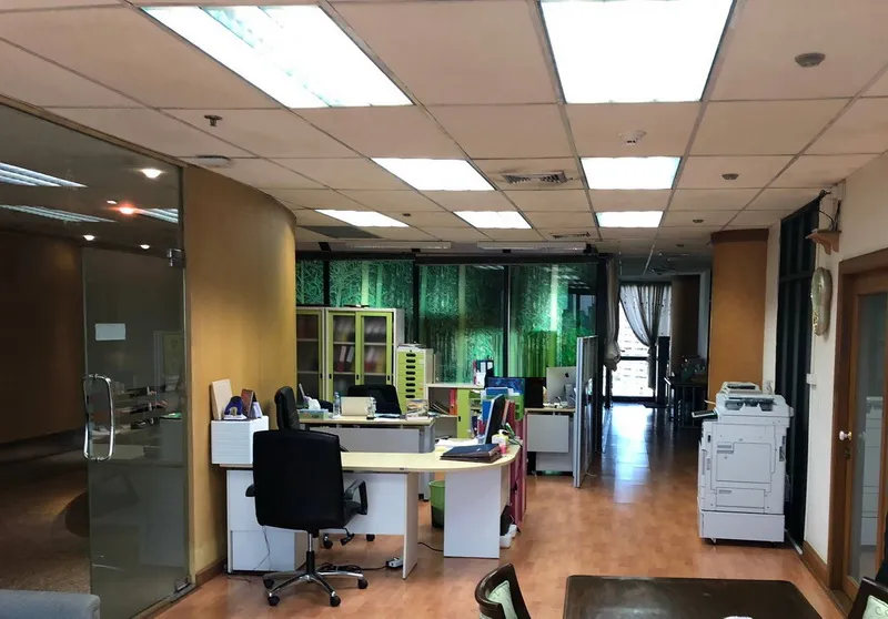 Open-plan office interior with desks, chairs, and office equipment, featuring a glass wall and greenery outside.