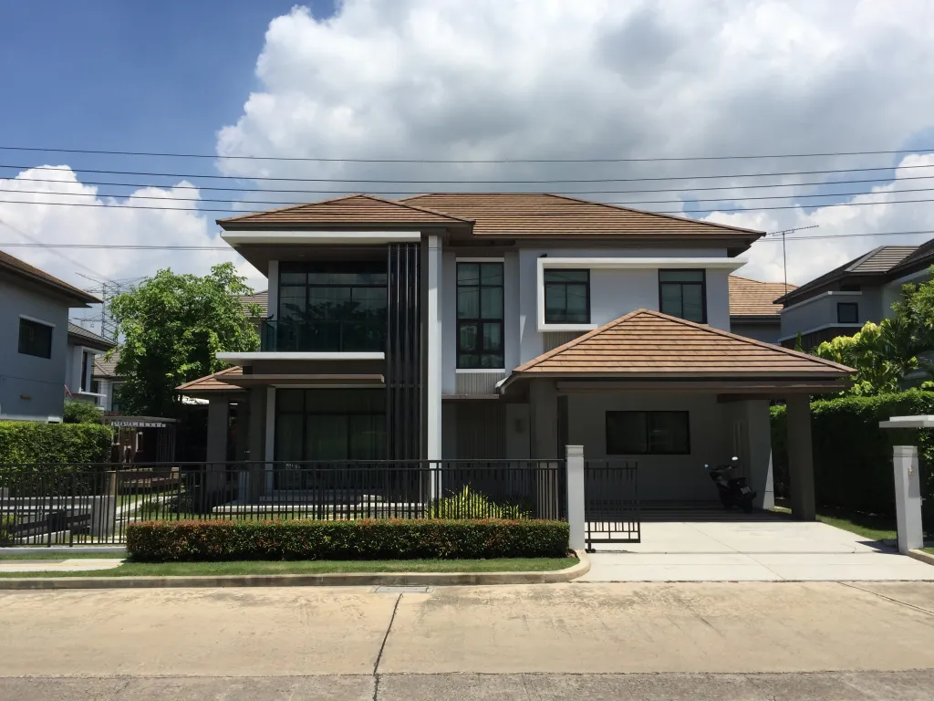 Two-story modern house with a pitched roof, large windows, and a driveway. Surrounded by greenery and a fence, under a partly cloudy sky.