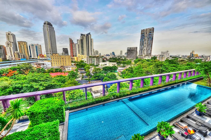 Rooftop infinity pool with modern city skyline and lush greenery in the background under a partly cloudy sky.