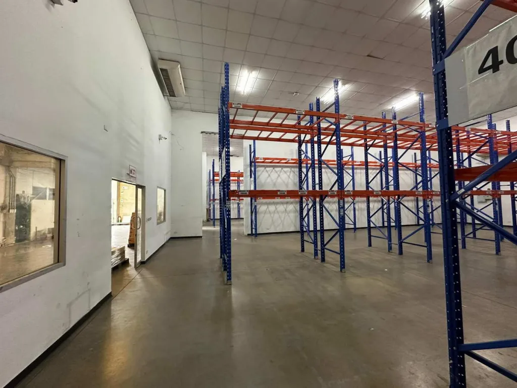 Empty warehouse interior with tall blue and orange shelving units, concrete floors, and fluorescent lighting.