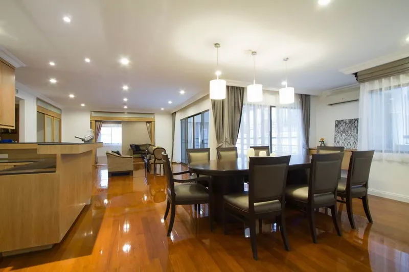 Spacious dining room with a dark round table and chairs, modern lighting, wooden floors, and large windows with curtains.