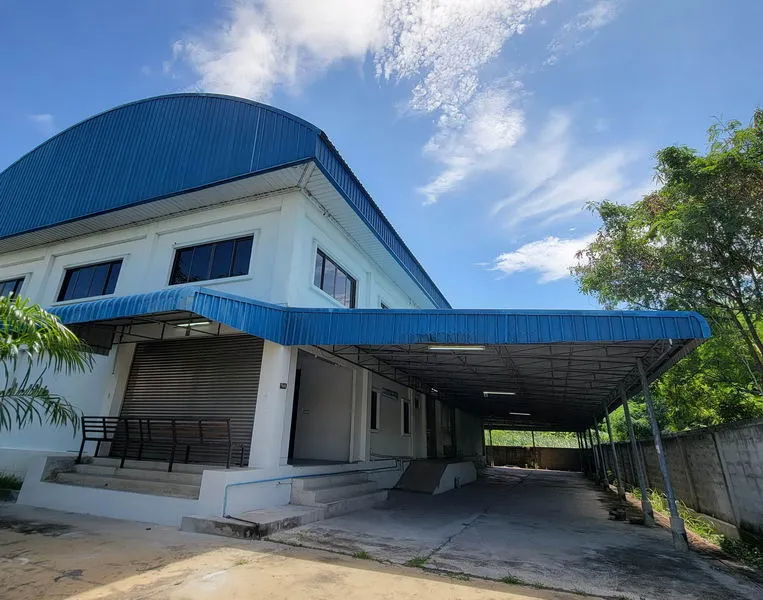 A large white building with a blue roof and awning, featuring a loading dock and driveway, surrounded by trees under a blue sky with clouds.