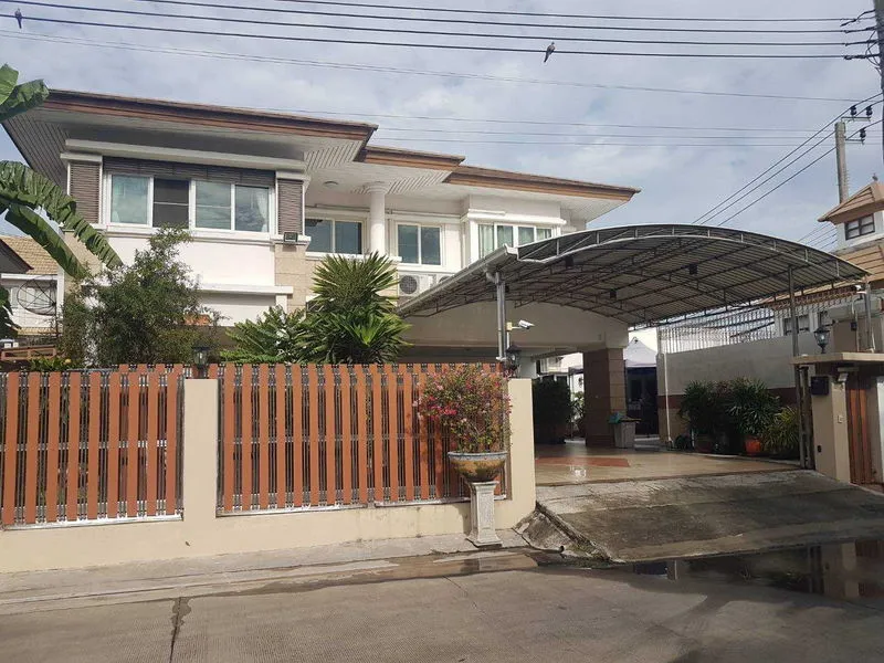 บ้านสองชั้น มีทางเข้าบ้านพร้อมประตูรั้ว โรงจอดรถโลหะ และต้นไม้เขียวชอุ่มอยู่ด้านหลังร