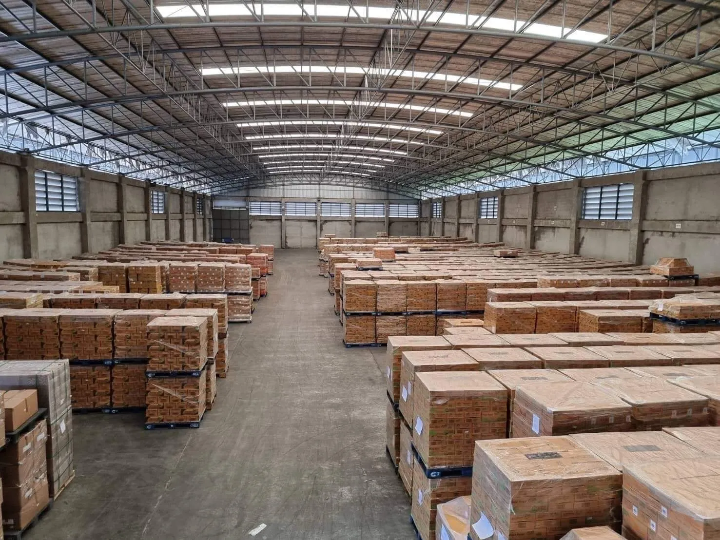 Warehouse interior with stacks of cardboard boxes arranged on pallets, under a high arched roof with metal beams.
