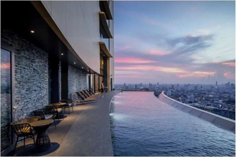 Rooftop infinity pool overlooking a city skyline at sunset, with lounge chairs and tables along a stone wall.