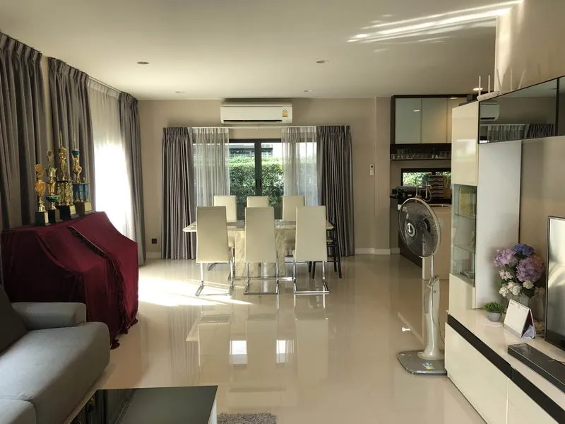 A modern dining room with a glass table, white chairs, curtains, and an air conditioner. A piano covered with a red cloth is on the left, and a fan and TV stand are on the right.