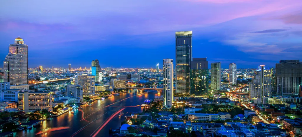 City skyline at dusk with illuminated buildings and a river reflecting lights under a twilight sky.