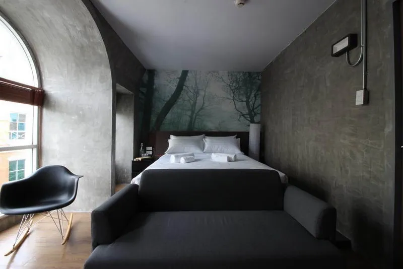 Modern bedroom with a forest mural, featuring a bed, gray sofa, black chair, and curved window.