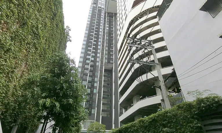 อาคารสูงทันสมัยที่มีหน้าต่างกระจกและอาคารสีขาวที่อยู่ใกล้เคียง ล้อมรอบด้วยต้นไม้และพ