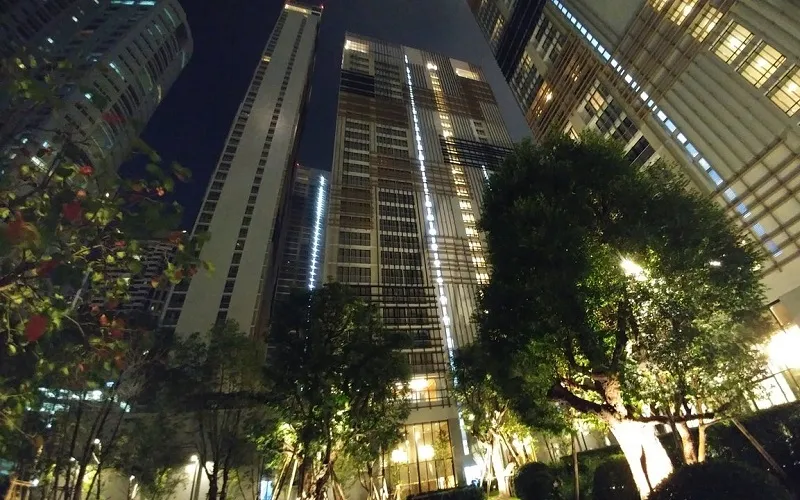 Skyscrapers with illuminated windows at night, surrounded by trees and lights in an urban setting.