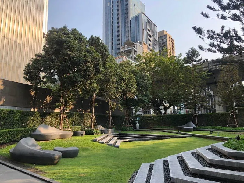 Urban park with modern stone benches, green lawn, trees, and skyscrapers in the background on a sunny day.