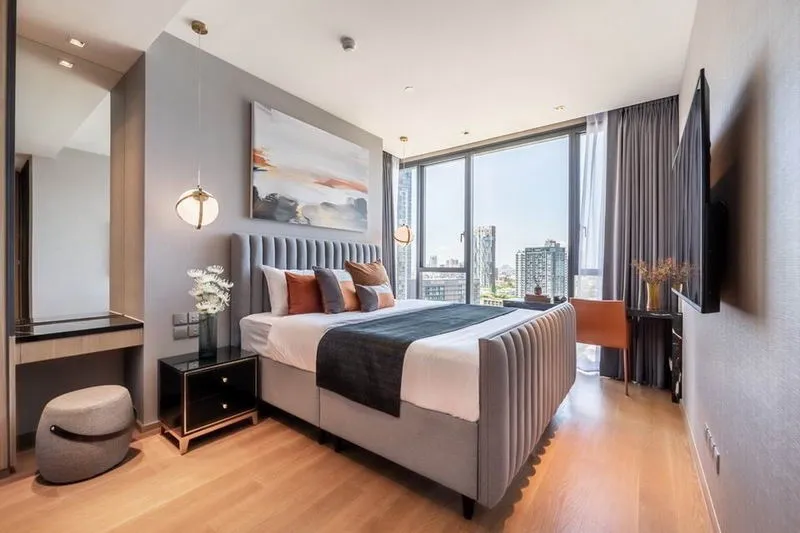 Modern bedroom with a large bed, art on the wall, pendant lighting, large windows with city view, and a small workspace with a chair and desk.