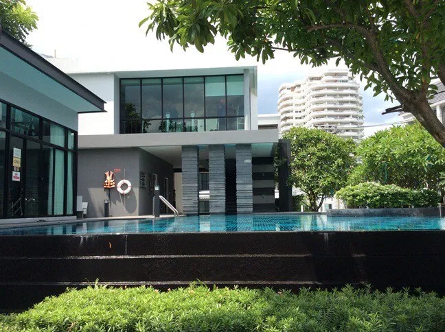 Modern house with large windows, surrounded by greenery and featuring a swimming pool. Nearby are high-rise buildings partially visible in the background.