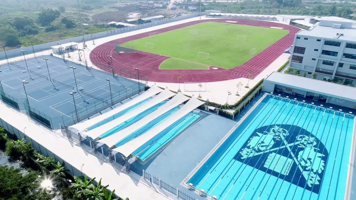 Aerial view of sports facilities including tennis courts, a swimming pool with a logo, and a grass field with a running track surrounding it.