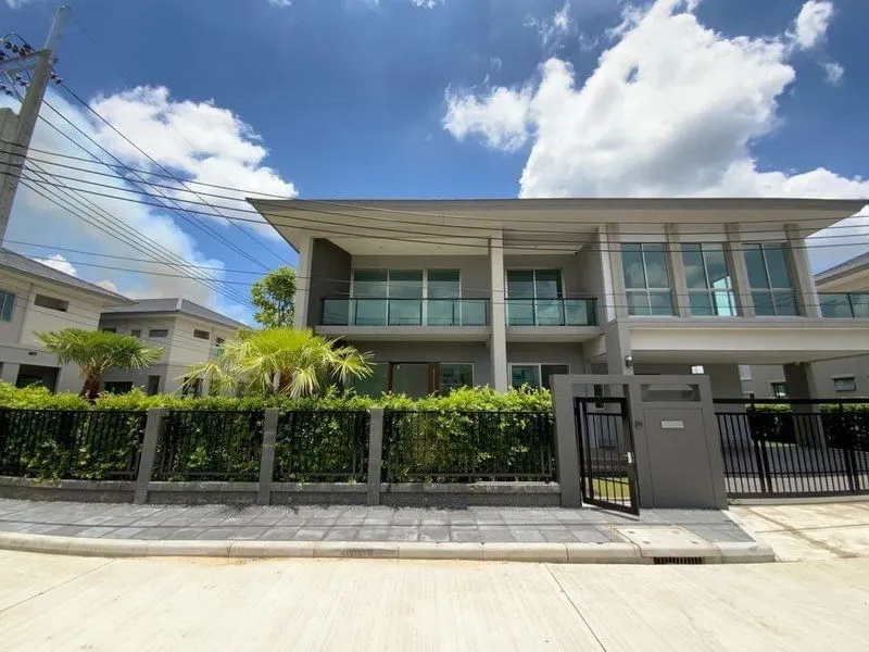 Modern two-story house with glass balconies, surrounded by greenery and a black gate, under a blue sky with clouds.